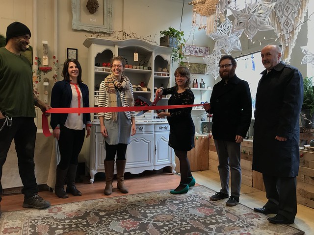 Mayor Patrick Madden, owner Sarah Bowen and other local representatives pose for a photo during the ribbon cutting of Twin Pots pottery studio. Participants are holding a red ribbon, with Bowen holding a large pair of scissors poised to cut the ribbon in half